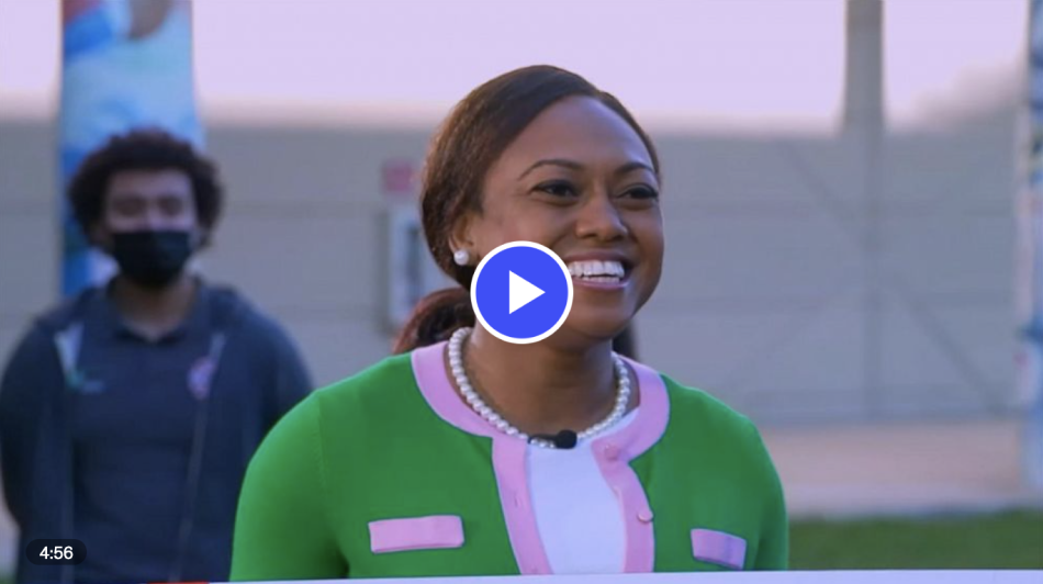 Teacher in green sweater smiles brightly, students stand behind her with a mask on