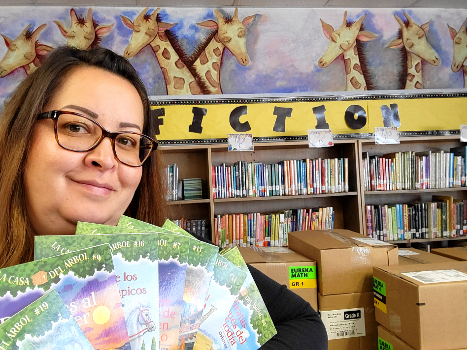 Photo of Mrs. Diaz holding up several books written in Spanish. Behind Mrs. Diaz is a stack of boxes and library shelves full of books